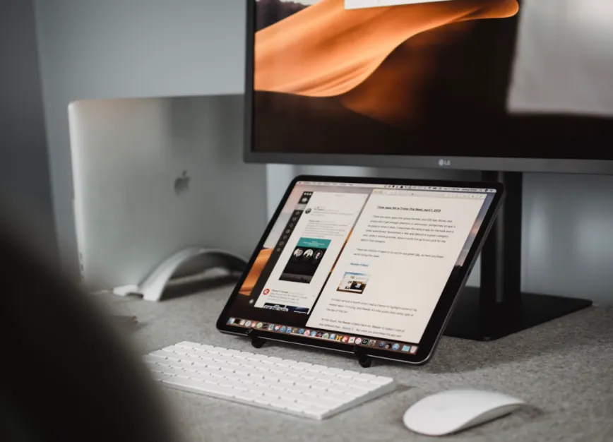 Bureau à domicile avec une tablette affichant un document au premier plan, un clavier et une souris devant elle, et un ordinateur portable et un écran externe à l'arrière-plan.